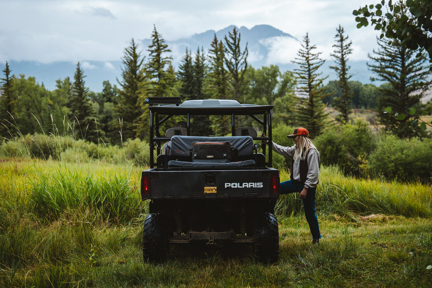 Woman standing next to a Polaris vehicle in a forested area with mountains in the background